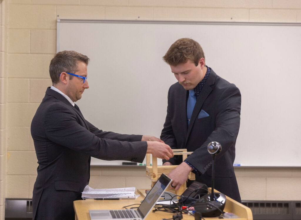 Photo of UW-Stout engineering students Luke Mertens, right, and Zachary Morgan demonstrating a wooden model of a machine they engineered for Ashley Furniture as part of their capstone course.